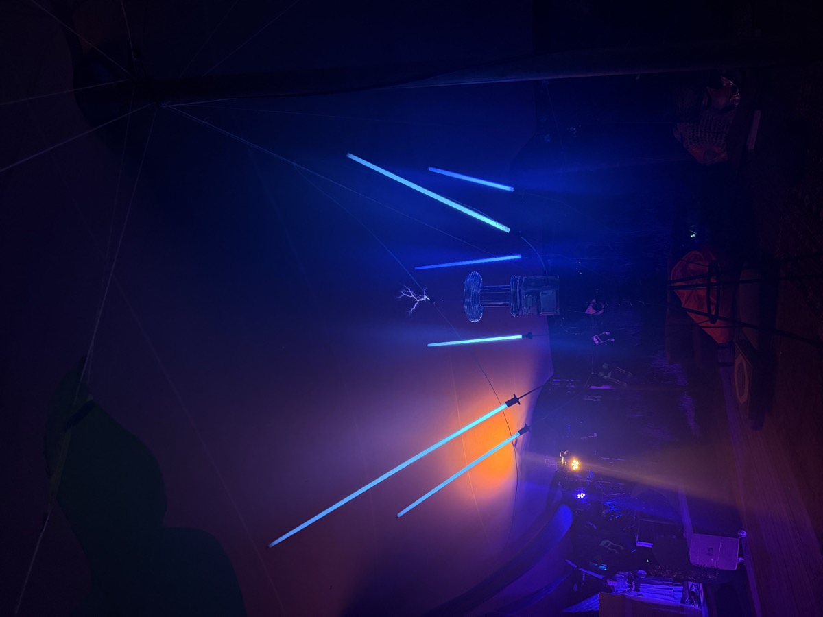 The Tesla coil arcs blue lightning; glowing tubes radiate around it in the tent