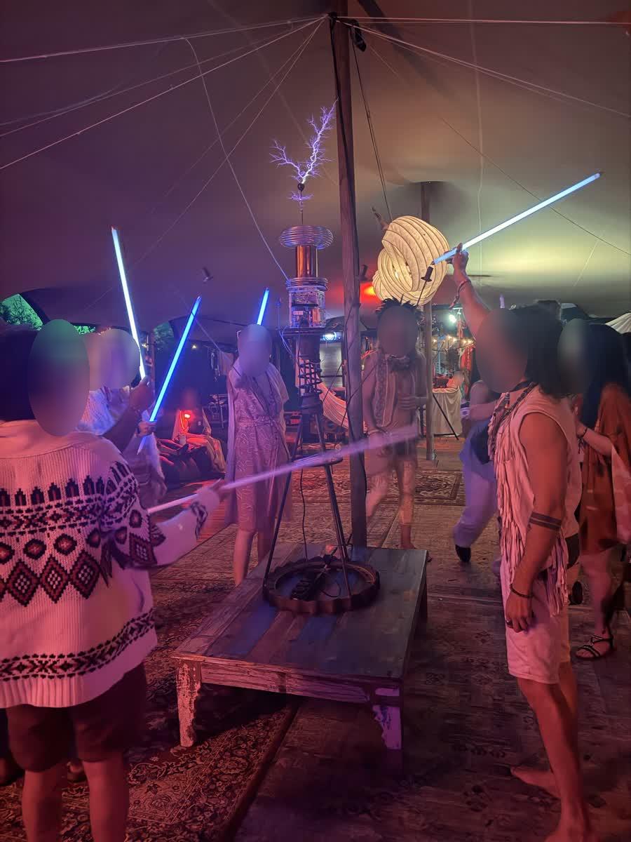 A crowd raises glowing tubes toward an arcing Tesla coil on a wooden plinth in a draped tent
