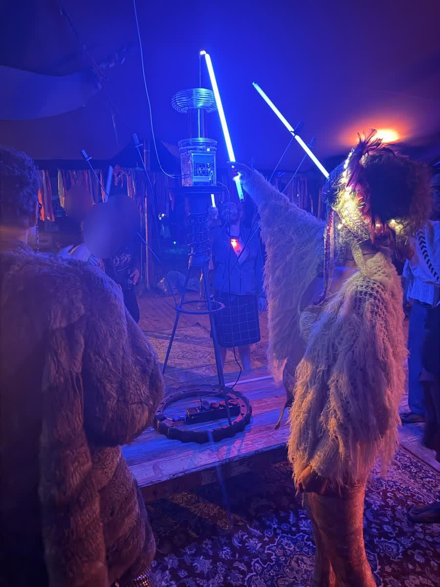 Figures in fur and feathers near the coil, tubes glowing blue in the deep tent light