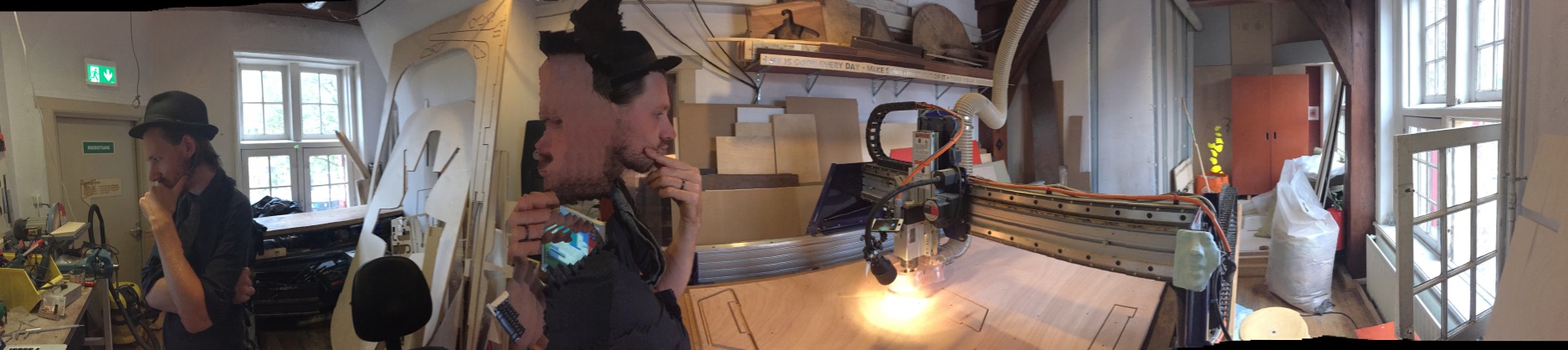 Spif at a shared Amsterdam workshop, panoramic — wearing a top hat, working at a CNC machine, doubled by the panorama stitch artifact
