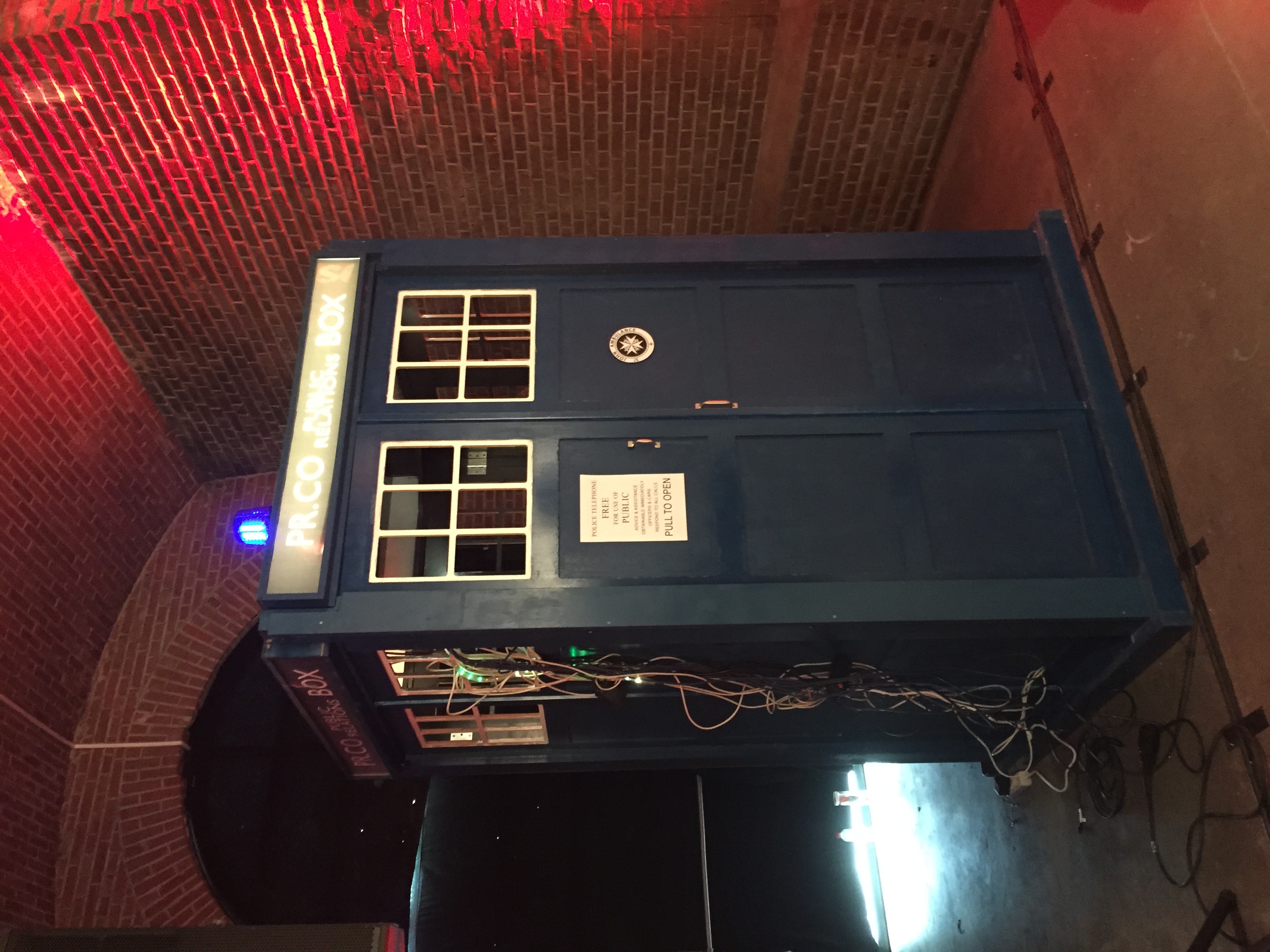 The TARDIS — life-sized blue police box recreation in a brick-vaulted hall, red lighting overhead, signage reading 'PR.CO PUBLIC BOX' and 'POLICE TELEPHONE FREE FOR USE OF PUBLIC'
