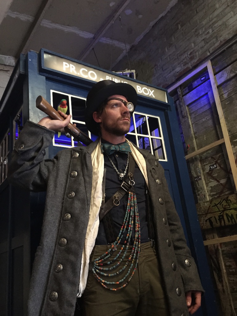 Spif at the Tardis — top hat with monocle, long grey wool coat, beaded necklaces, holding a wooden tool, Tardis police box behind